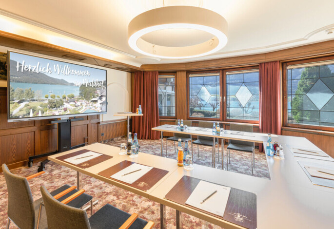 Conference room at Waldhof Fuschlsee Resort with tables, chairs, and a screen displaying "Welcome to Waldhof." Large windows offer a lake view.