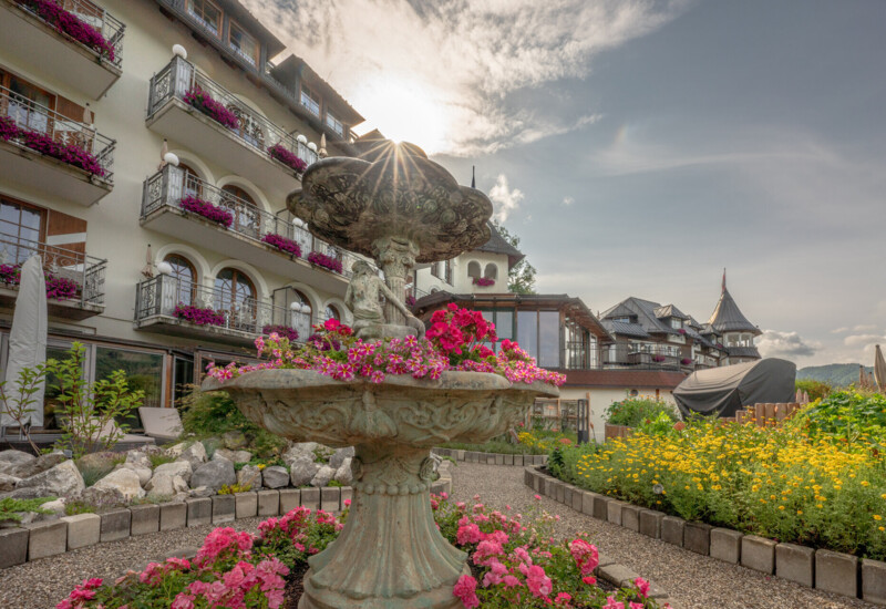 Ein malerischer Brunnen mit Blumen vor dem Waldhof Fuschlsee Resort mit Balkonen, die mit Pflanzen geschmückt sind, unter einem strahlenden Himmel.