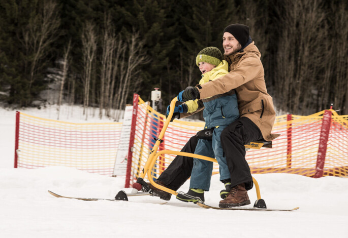 Father and child enjoying a snowbike ride on a snowy slope with safety netting and forest backdrop.