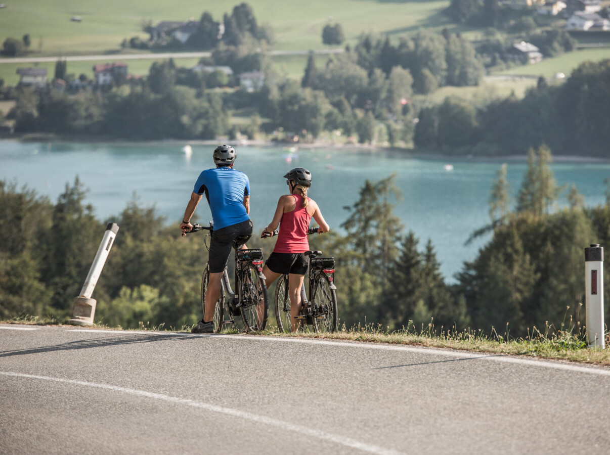 Two cyclists pause to enjoy a scenic view of lake Fuschl surrounded by lush greenery.