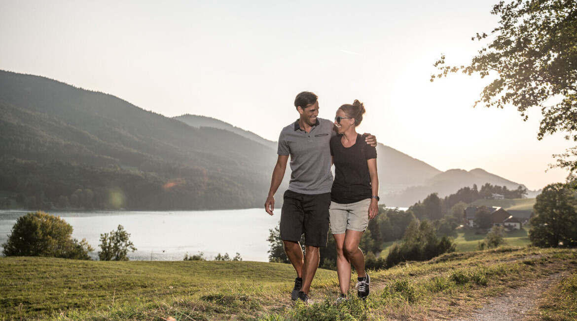 Couple walking on a scenic lakeside path with mountains in the background, enjoying a sunny day.