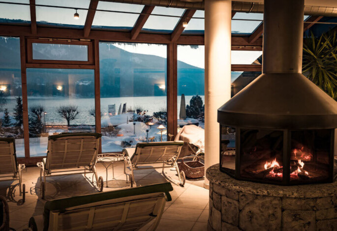 Cozy indoor area with fireplace and chairs overlooking a snowy, scenic lake view through large windows.