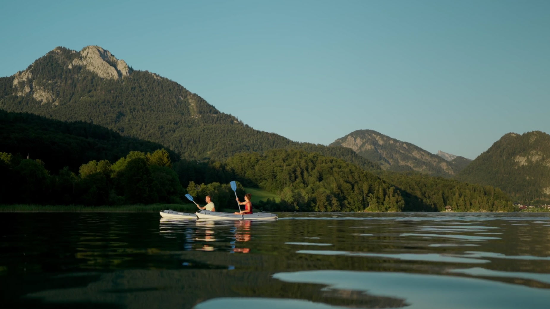 Two people kayaking on a calm lake surrounded by lush forest and scenic mountains under a clear sky.