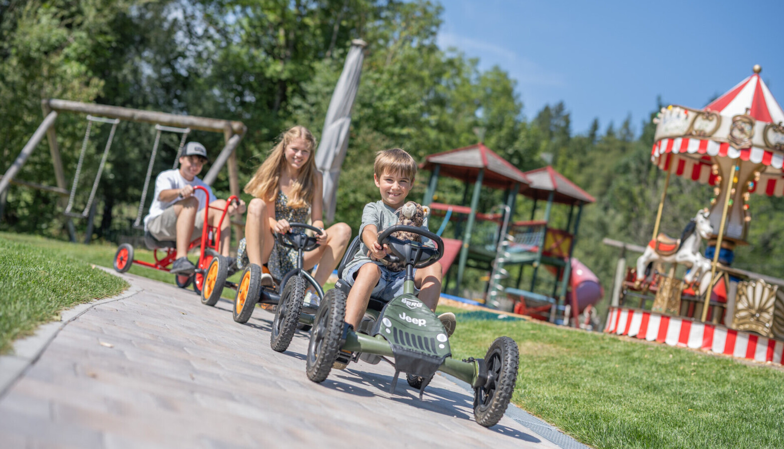 Children enjoying go-karting at Waldhof Fuschlsee Resort with a carousel and playground in the background.