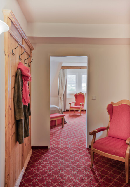 A cozy hotel room with red patterned carpet, wooden furniture, and a coat hanging on a rack.