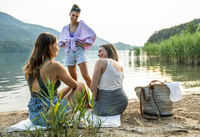 Three women enjoying a scenic lakeside picnic, surrounded by lush greenery, with a woven basket nearby.