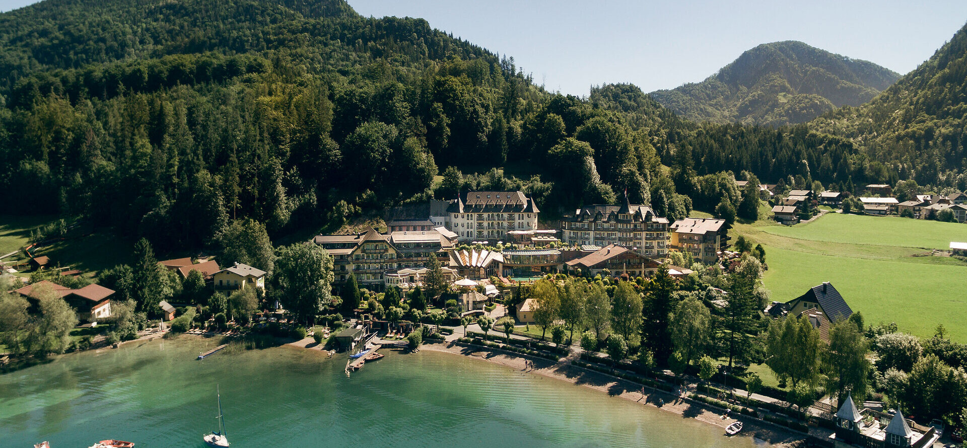 Aerial view of Waldhof Fuschlsee Resort by a tranquil lake, surrounded by forested hills and mountains.