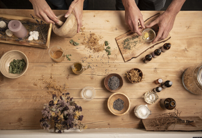 Hands preparing herbal mixtures on a wooden table with dried herbs, oils, and candles.