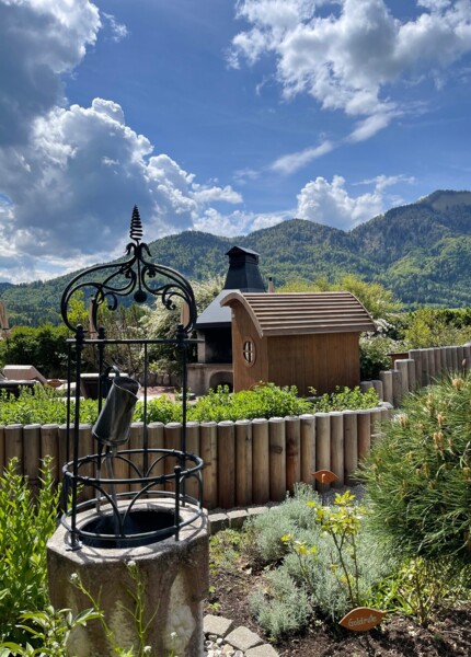 Scenic garden with a well, surrounded by lush greenery and mountains under a blue sky with clouds.