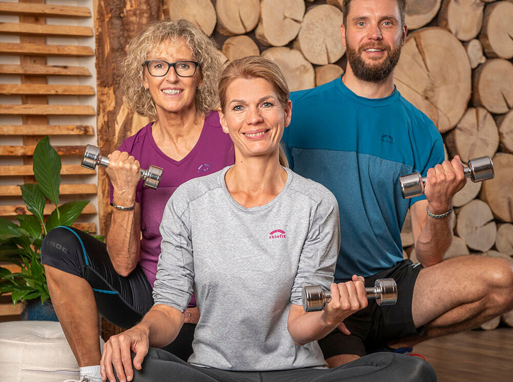 Three people sitting on exercise mats, holding dumbbells, and smiling, with a wood log background.