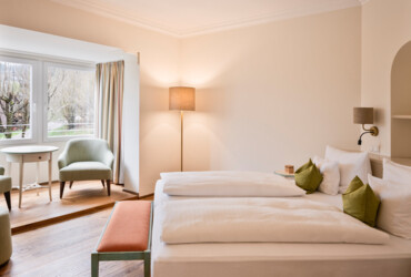 Bright hotel room with a cozy double bed, green pillows, seating area, and large windows overlooking trees.