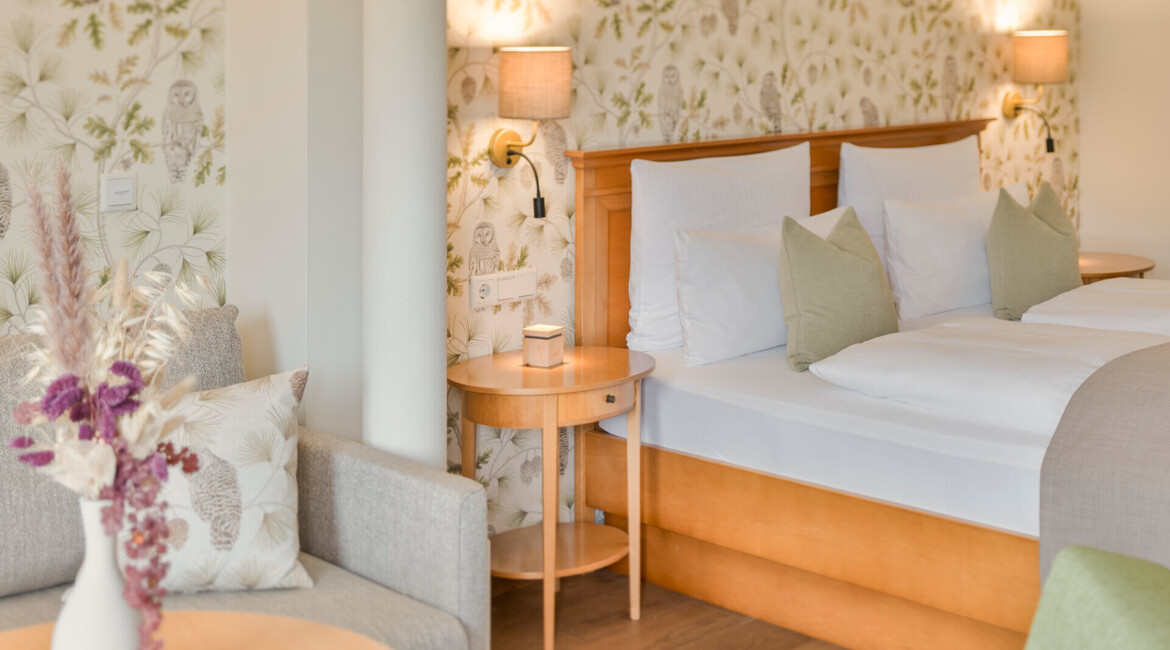 Hotel room with floral wallpaper, wooden furniture, and a vase of dried flowers.
