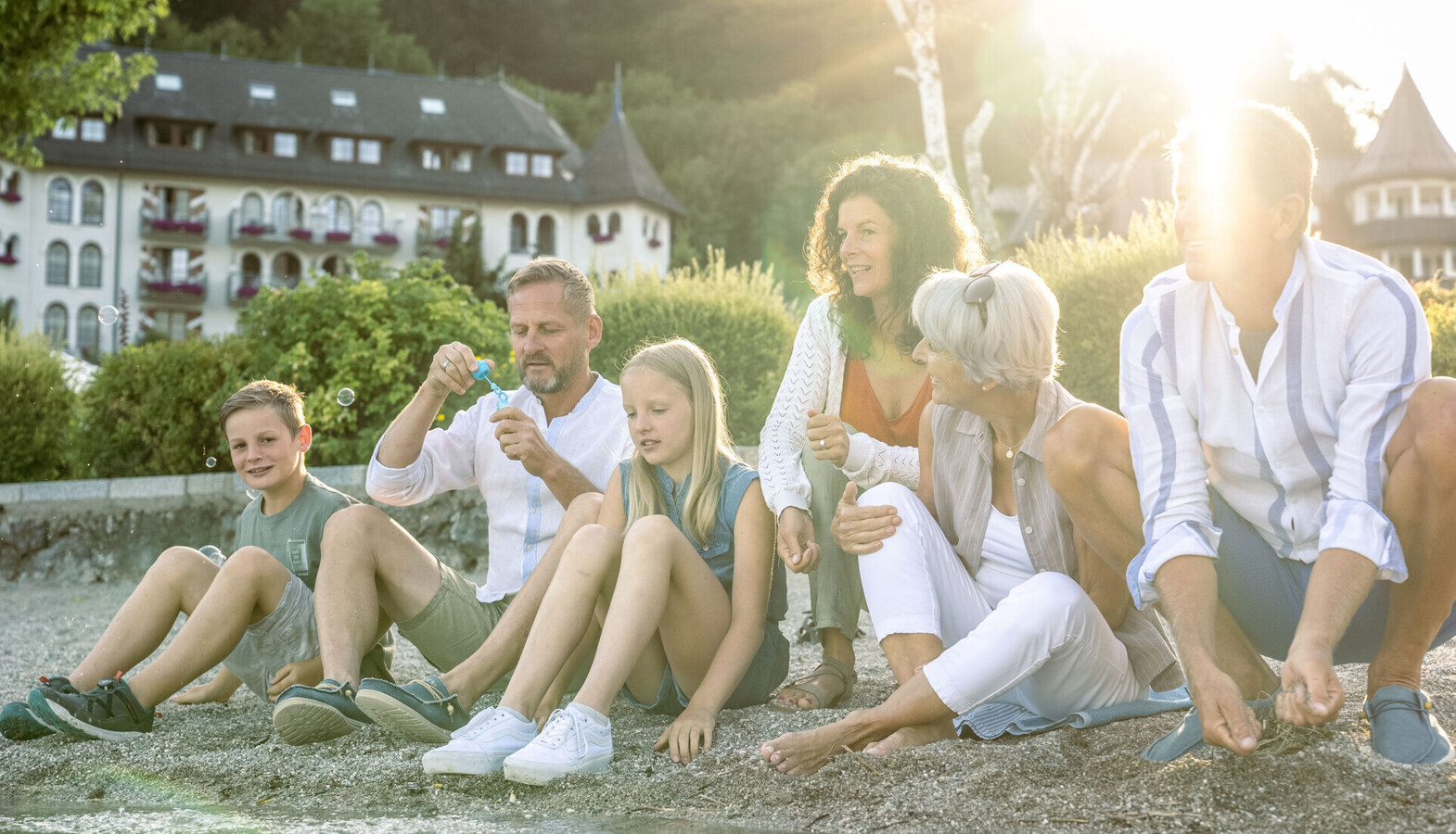 A family enjoying a sunny day by the water, with Waldhof Fuschlsee Resort and greenery in the background.