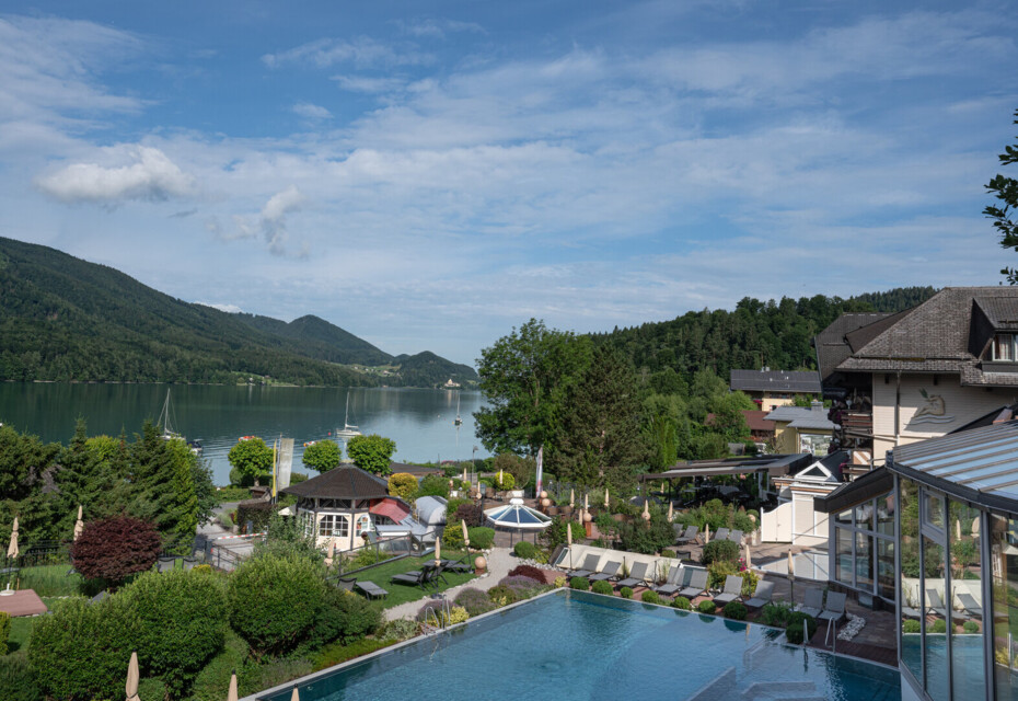 View of Waldhof Fuschlsee Resort with pool, garden, and lake surrounded by mountains under a blue sky.