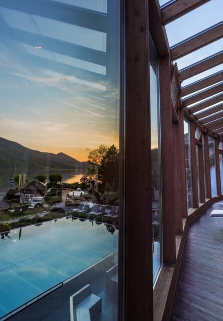 Glass-walled lounge with sun chairs overlooking a scenic lake and mountains at sunset.