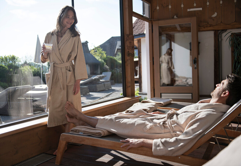 A woman and man in robes relax by a sunny window at a spa.