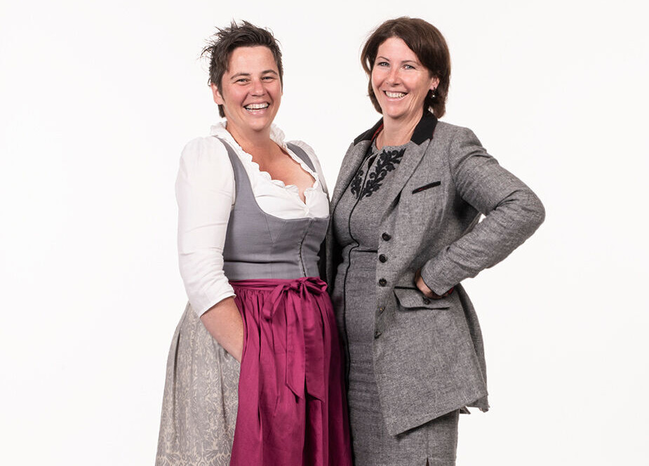 Two women smiling, one in traditional attire with a pink apron, and the other in a gray suit, standing together on a plain background.