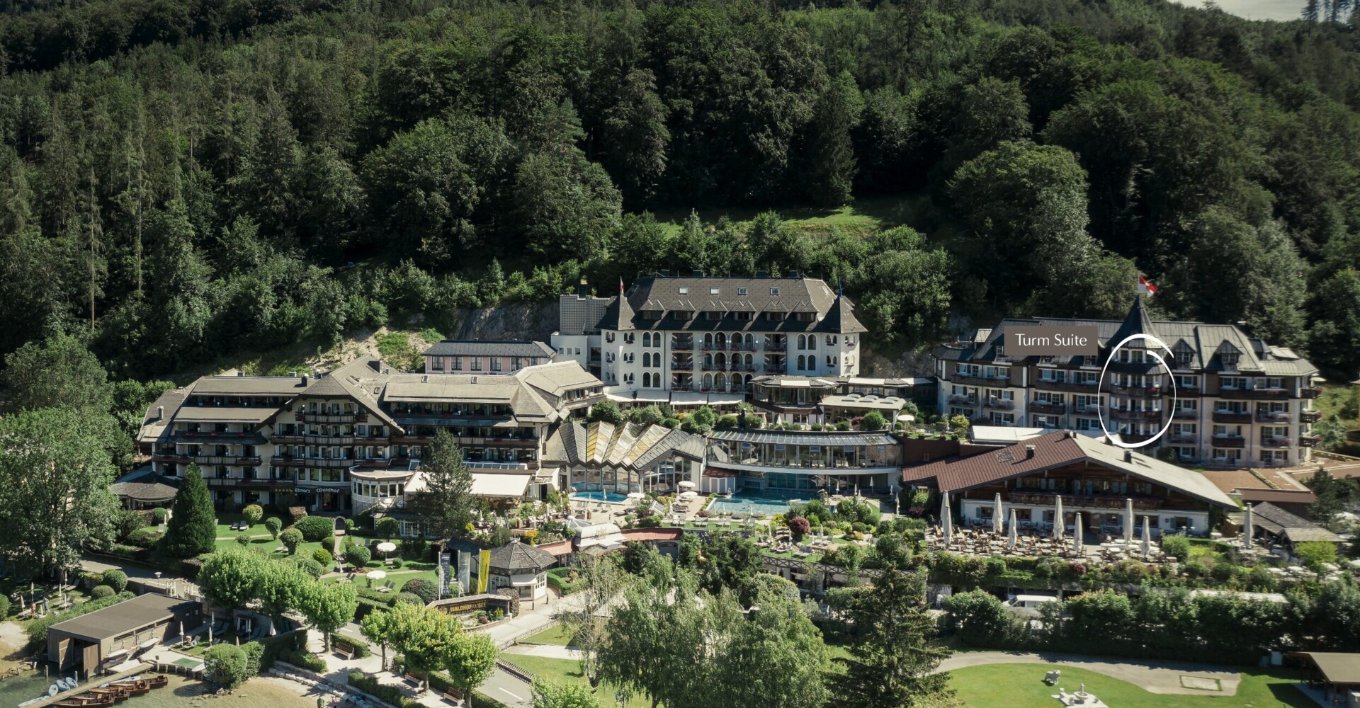 Aerial view of Waldhof Fuschlsee Resort surrounded by lush greenery, with highlighted Turm Suite, near a serene lakeside.