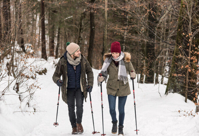 A couple enjoys a winter hike through a snowy forest, using trekking poles for support.