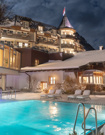 Snow-covered hotel with a glowing pool at night, surrounded by a scenic winter landscape.