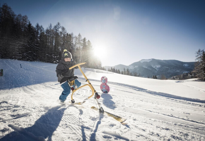 Child sledding on a snowy hill with a scenic mountain backdrop under a clear blue sky.