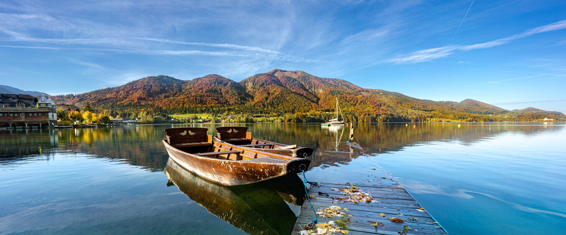 Ein Holzboot liegt an einem Steg auf einem ruhigen See, umgeben von herbstlichen Bergen unter klarem blauem Himmel.