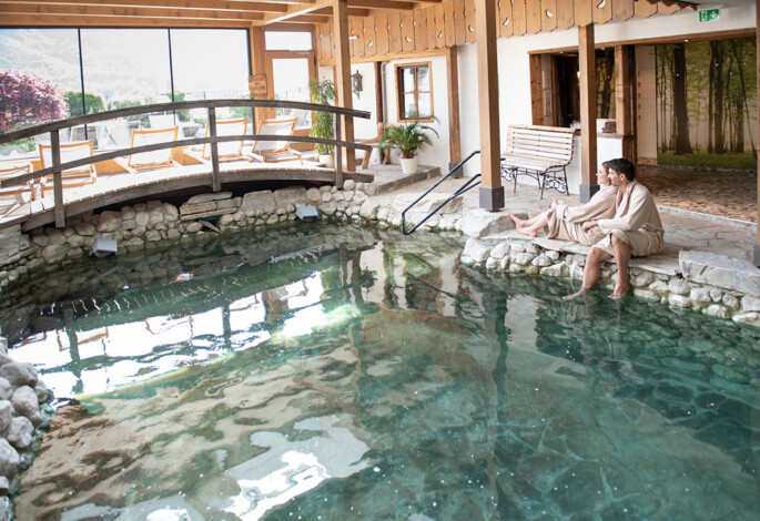 Couple in robes sitting by an indoor pool at Waldhof Fuschlsee Resort, with lounge chairs and stone decor.