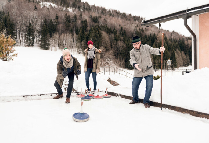 People playing ice stock sport outdoors in winter, surrounded by snowy trees and mountains.