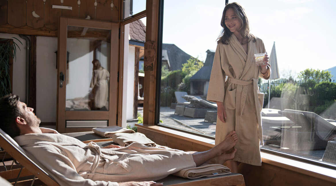 A man and woman relax in robes by a large window at Waldhof Fuschlsee Resort, enjoying a serene indoor setting.