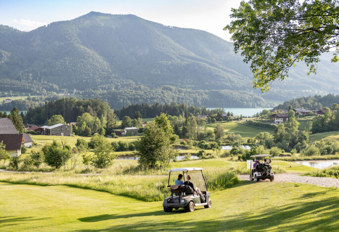 Golf carts on a lush, scenic course with mountains and a lake in the background.