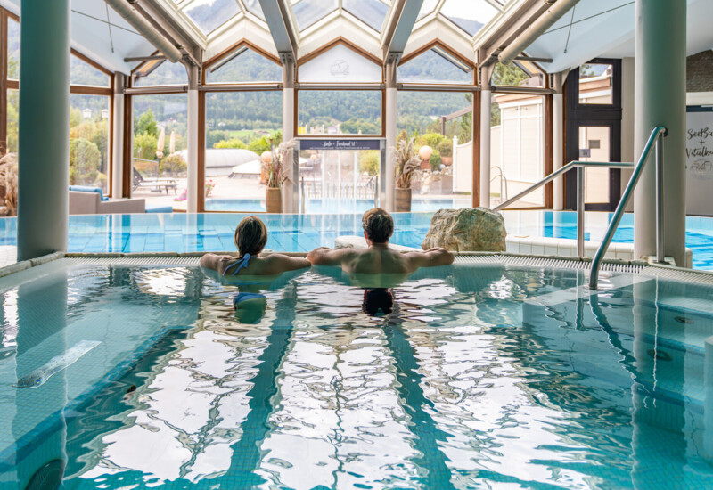 Couple relaxing in an indoor pool with scenic mountain views through large windows.