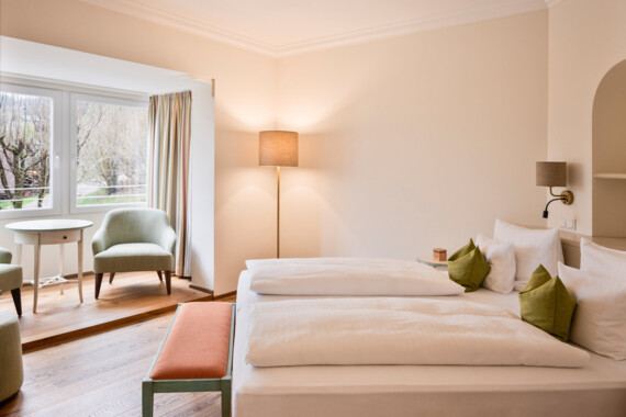 Bright hotel room with a cozy double bed, green pillows, seating area, and large windows overlooking trees.