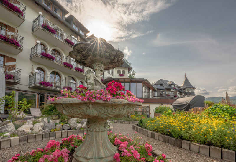 Fountain surrounded by vibrant flowers in the garden of Waldhof Fuschlsee Resort, with the hotel building in the background under a sunny sky.