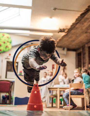 A child jumps through a hoop during an indoor activity at a play center, with other children watching in the background.