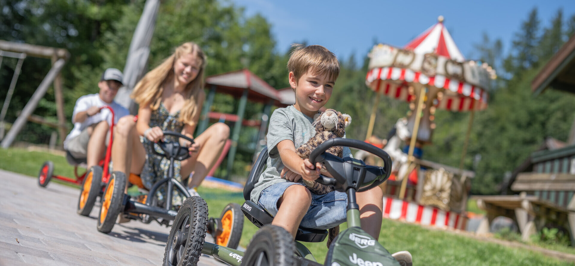 Children enjoying pedal go-karts at Waldhof Fuschlsee Resort, with a carousel in the background.