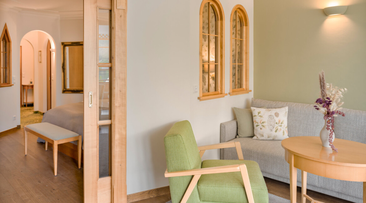 Cozy room with wooden furniture, green armchair, and beige sofa. Sliding door opens to a bedroom. Soft lighting creates a warm ambiance.