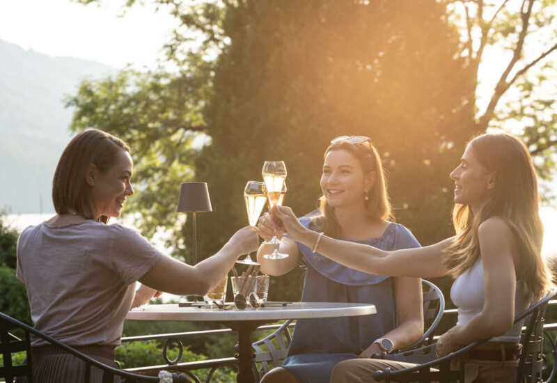 Three women toast with drinks on a scenic terrace surrounded by lush greenery.