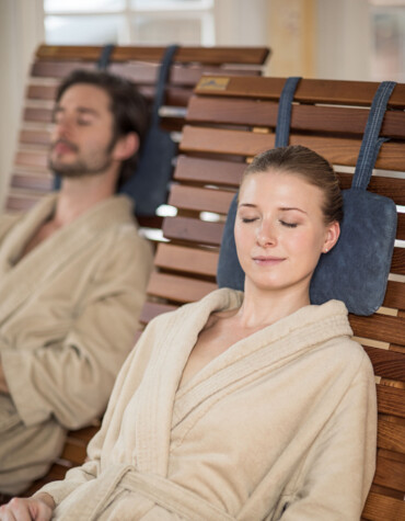 Zwei Personen entspannen in Bademänteln auf Holzliegen in einem Spa-Bereich.