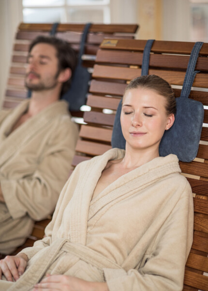 Two people relaxing on wooden lounge chairs, wearing bathrobes, with eyes closed in a peaceful setting.