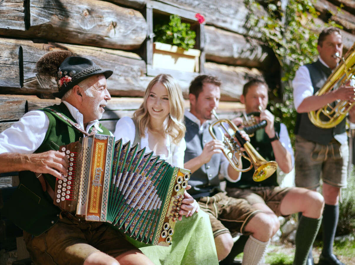 A group in traditional attire plays folk music with accordion and brass instruments, sitting by a rustic wooden building.