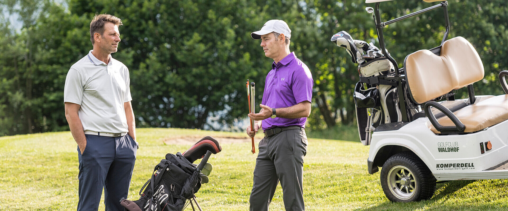 Two men talking on a golf course near a cart, one holding a club.