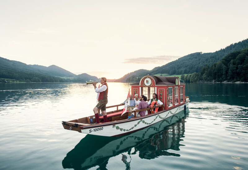 A boat with people in traditional attire sails on a serene lake surrounded by lush, scenic mountains at sunset.