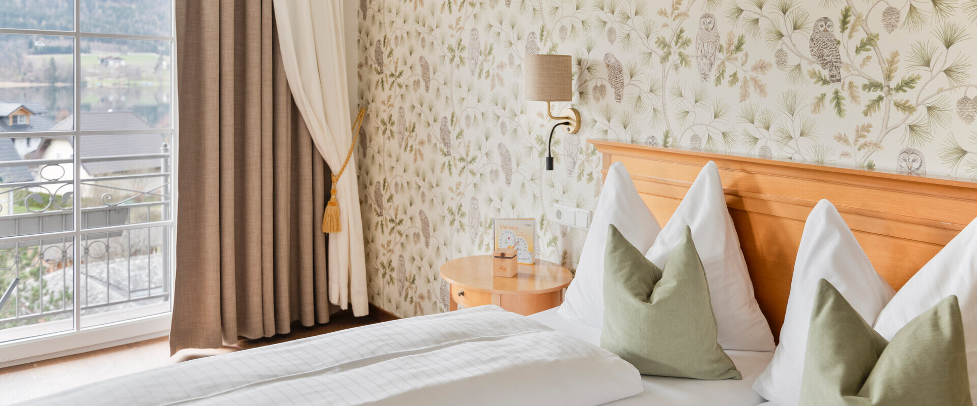 Hotel room with floral and owl wallpaper, a double bed with green pillows, and a scenic view of the lake and mountains through a large window.