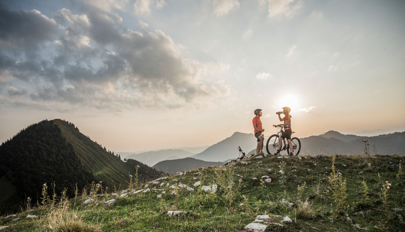 Zwei Radfahrer genießen den Sonnenuntergang auf einem Berggipfel mit Blick auf bewaldete Hügel und eine malerische Landschaft.