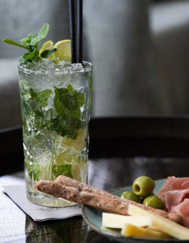 Ein erfrischender Cocktail mit Minze und Limette neben einem Teller mit Oliven, Prosciutto und Käse auf einem Tisch.
