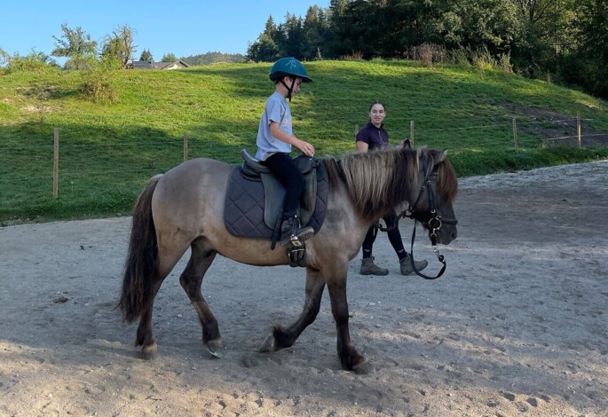 Kind reitet auf einem Pferd in grüner Landschaft unter blauem Himmel, begleitet von einem Erwachsenen.