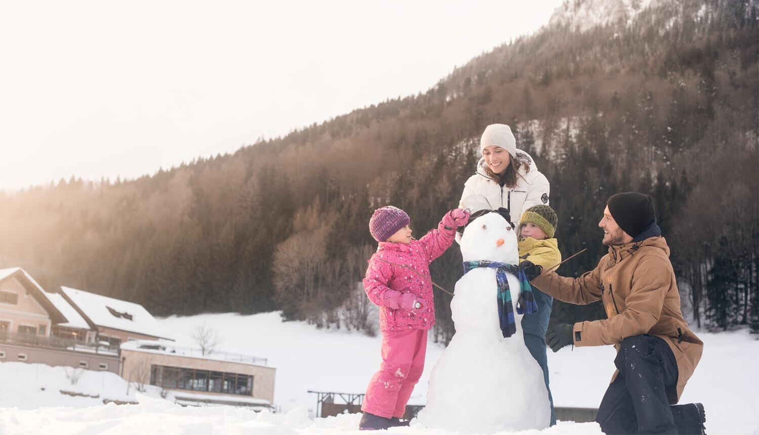 A family building a snowman in a snowy landscape with mountains in the background, enjoying a winter day.