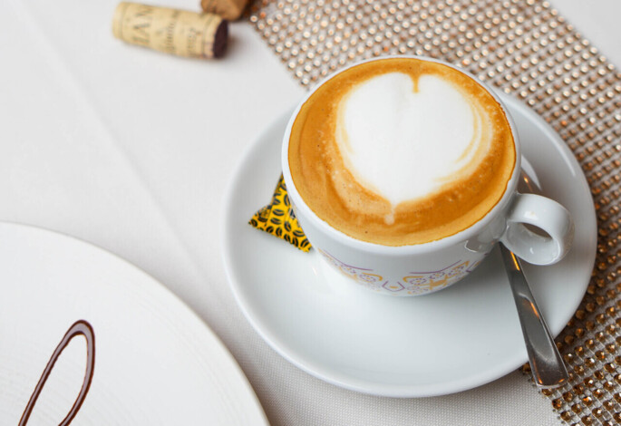 A cappuccino with heart-shaped foam on a table with a candle and wine corks.