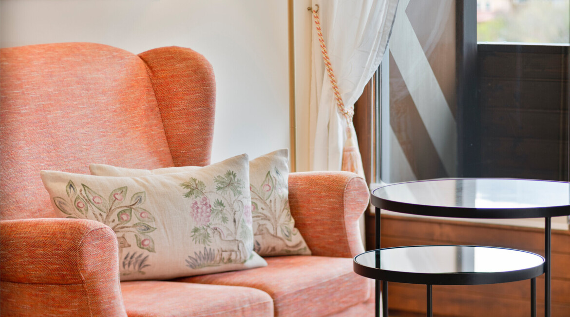 Cozy seating area with an orange armchair, decorative pillows, and a standing lamp, set near a window with a scenic view.