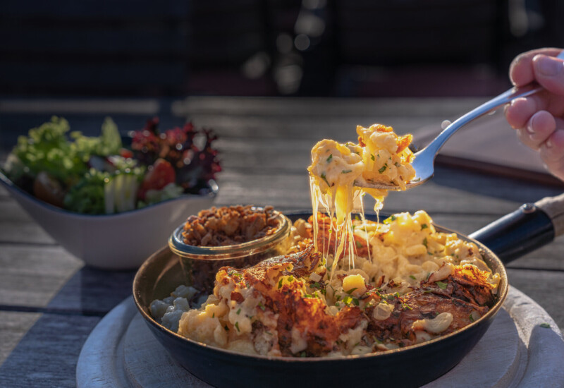 Käsespätzle mit geschmolzenem Käse im Pfännchen, serviert mit einem frischen Salat im Hintergrund auf einem Tisch.